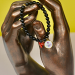 Bracelet Madonuccia noir à 2 pampilles en perles de verre noires, orné d’un fragment de corail naturel et d’une médaille représentant la Madonuccia, symbole de la tradition corse et de la culture méditerranéenne.